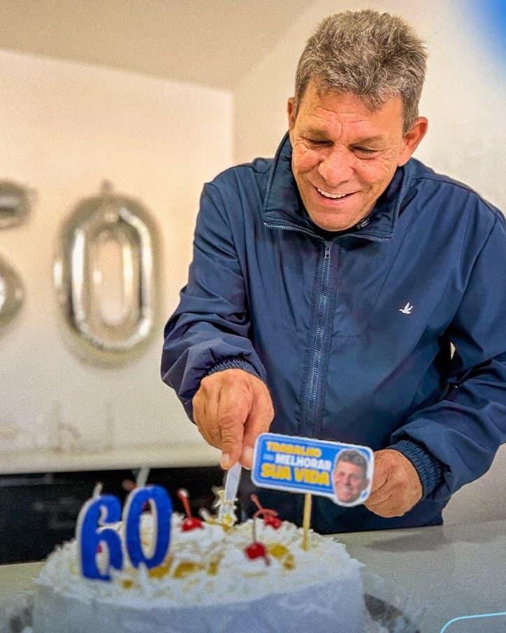 Carlos Russo sorri ao cortar o bolo de 60 anos, com velas e balões comemorativos